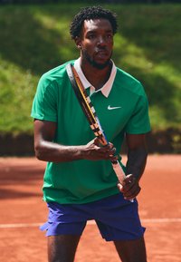 Meesatleet rohelises lühikeste varrukatega särgis, millel on valge kaelus, ja sinistes šortse, hoiab tennisereketit saviväljakul.