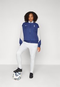 Blå och grå zip-jacka med logotyp, tillsammans med vita leggings. Svarta gympaskor. En fotboll placerad under foten.