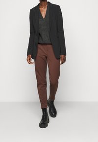 Schwarz maßgeschneiderte Blazer über einem grauen V-Ausschnitt-Pullover, kombiniert mit braunen Slim-Fit-Hosen und schwarzen Schnürstiefeln. Glatte Texturen und moderne Passform.