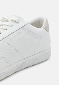 Baskets en cuir blanc à finition texturée, avec des accents en daim gris, des lacets plats et un logo embossé sur le côté. Semelle en caoutchouc perforée.