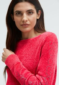 Junge Frau mit langen braunen Haaren, die einen leuchtend roten strukturierten Pullover mit metallischem Text und Strassdetails an den Ärmeln trägt.