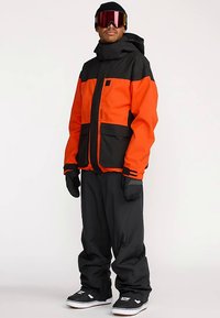 Veste de ski orange et noire avec fermeture éclair frontale, grandes poches et capuche ajustable. Associée à un pantalon noir ample et des chaussures de neige.