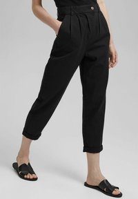 Pantalon noir taille haute avec plis et poches latérales, jambe fuselée, ourlets retroussés, associé à des sandales noires à enfiler. Tissu en coton lisse.