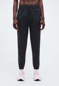 Άτομο που φοράει μαύρο ελαστικό παντελόνι jogger και ανοιχτό ροζ αθλητικά παπούτσια, στέκεται σε απλό ανοιχτό γκρι φόντο.