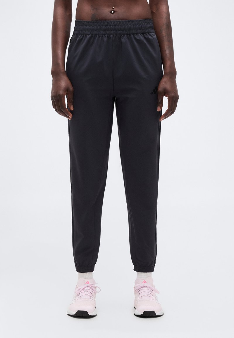 Άτομο που φοράει μαύρο ελαστικό παντελόνι jogger και ανοιχτό ροζ αθλητικά παπούτσια, στέκεται σε απλό ανοιχτό γκρι φόντο.