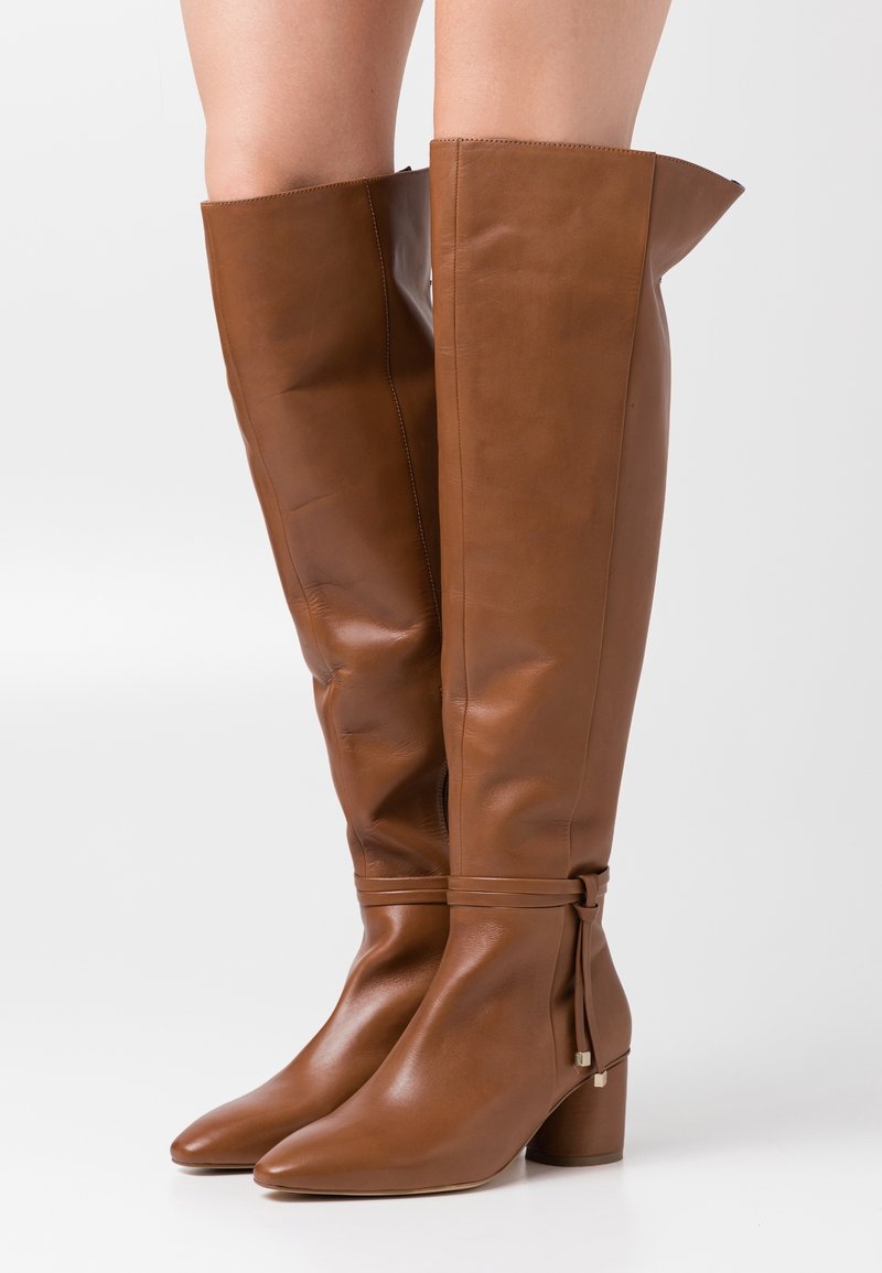 Bottes en cuir marron montant jusqu'au genou, avec un bout pointu, une finition texturée et un talon carré. Elles présentent un détail froncé sous le genou.