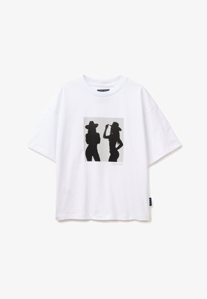 T-shirt bianca con sagome nere di due donne che indossano cappelli a tesa larga, una con le braccia incrociate e l'altra che tiene il cappello.