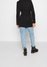 Abrigo negro con cinturón, de corte ajustado, combinado con jeans de color azul claro y botas negras con cordones. Tejido suave y diseño de longitud media.