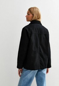 Svart bomullsjacka med krage, med rak passform och en yoke-seam bak. Modellen bär ljusblå denimjeans.