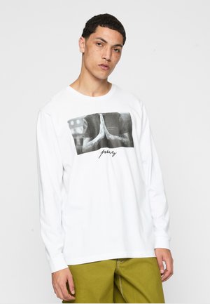 Jeune homme avec des cheveux courts et bouclés portant une chemise blanche à manches longues avec un imprimé mains en prière noir et blanc, et un pantalon vert.