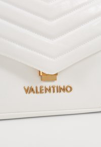 Vitt läderhandväska med en quiltad chevronmönster, gyllene detaljer och varumärket "VALENTINO" präglat i guld.
