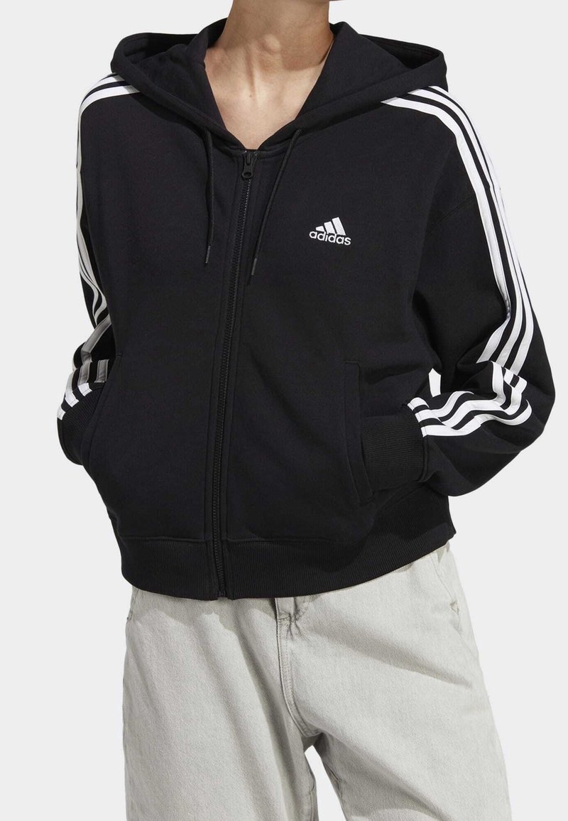 Person som bär en svart Adidas zip-up hoodie med vita ränder på ärmarna och logotypen på bröstet, ihop med ljusa byxor.