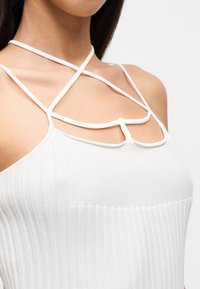 Κοντινό πλάνο γυναίκας που φοράει λευκό ριμπ μπλουζάκι χωρίς μανίκια με λεπτούς, σταυρωτούς ιμάντες στο πάνω μέρος του στήθους και στους ώμους.