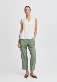Top bianco senza maniche con scollo a V e dettagli in pizzo, abbinato a pantaloni larghi di un leggero verde. Ballerine marroni completano l'outfit.