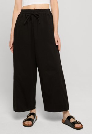 Nő laza, fekete, széles szárú nadrágot visel, rugalmas derékpánttal és húzózsinórral, fekete papucsokkal, amelyeken csatok vannak.