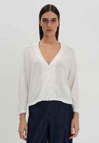 White cardigans with a V-neck, long sleeves, ribbed texture, and button closure, paired with dark blue pants. Smooth fabric with a relaxed fit.