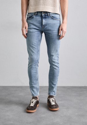 Osoba ubrana w jasnoniebieskie obcisłe dżinsy i ciemnobrązowe trampki z białymi sznurowadłami, stojąca na szarej podłodze przy jednolitej ścianie.