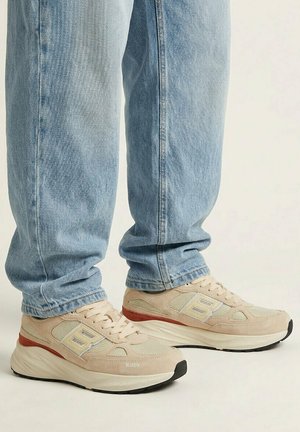 Persona con jeans azzurri chiari e sneaker beige con accenti rossi, in piedi su uno sfondo chiaro e uniforme.