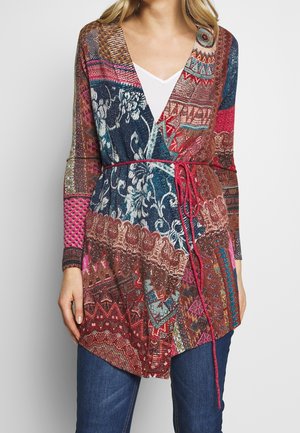 Femme portant un cardigan patchwork multicolore avec des motifs floraux et géométriques, attaché avec une fine ceinture rouge, par-dessus un top blanc et un jean bleu.