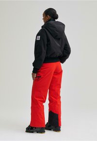 Fekete kapucnis dzseki rugalmas mandzsettákkal, élénk piros nadrággal, amely cipzáras zsebekkel és fekete részletekkel rendelkezik a bokáknál. Fekete lábbeli.