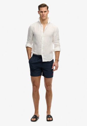 Uomo che indossa una camicia bianca di lino a maniche lunghe con polsini arrotolati, pantaloncini blu navy, sandali neri e un orologio da polso, in piedi su sfondo bianco.