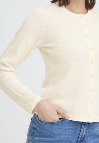 Cardigan beige clair avec un col rond et des manches longues, doté d'une fermeture à boutons sur le devant et de poignets côtelés. Texture lisse, coupe ajustée.
