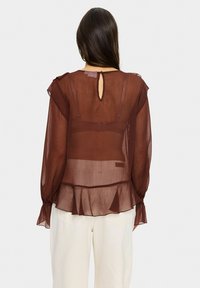 Brown sheer blouse with a ruffled hem, keyhole back, and subtle texture. Features long sleeves with elastic cuffs. Paired with light trousers.