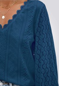 Blauw kanten top met een gegolfde V-halslijn, ingewikkelde oogjespatronen en gestructureerde lange mouwen, met een zachte stofafwerking.