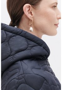Chaqueta acolchada de puffer en color navy con cuello alto, que presenta un pendiente de aro dorado con un gran colgante de perla redonda y texturizada.