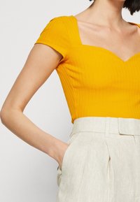 Gul ribbad topp med korta ärmar och fyrkantig halsringning, kombinerad med ljusbeiga linbyxor med sidfickor och veck.