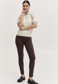 Beige gestricktes Poloshirt mit kurzen Puffärmeln, drei Knöpfen und einem gerippten Saum, kombiniert mit figurbetonten braunen Hosen und schwarzen Mokassins.