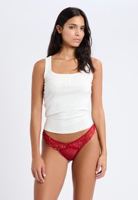 Top sin mangas de crema con escote cuadrado y detalles de botones, combinado con tangas de encaje rojo transparente que presentan lunares y patrones florales.
