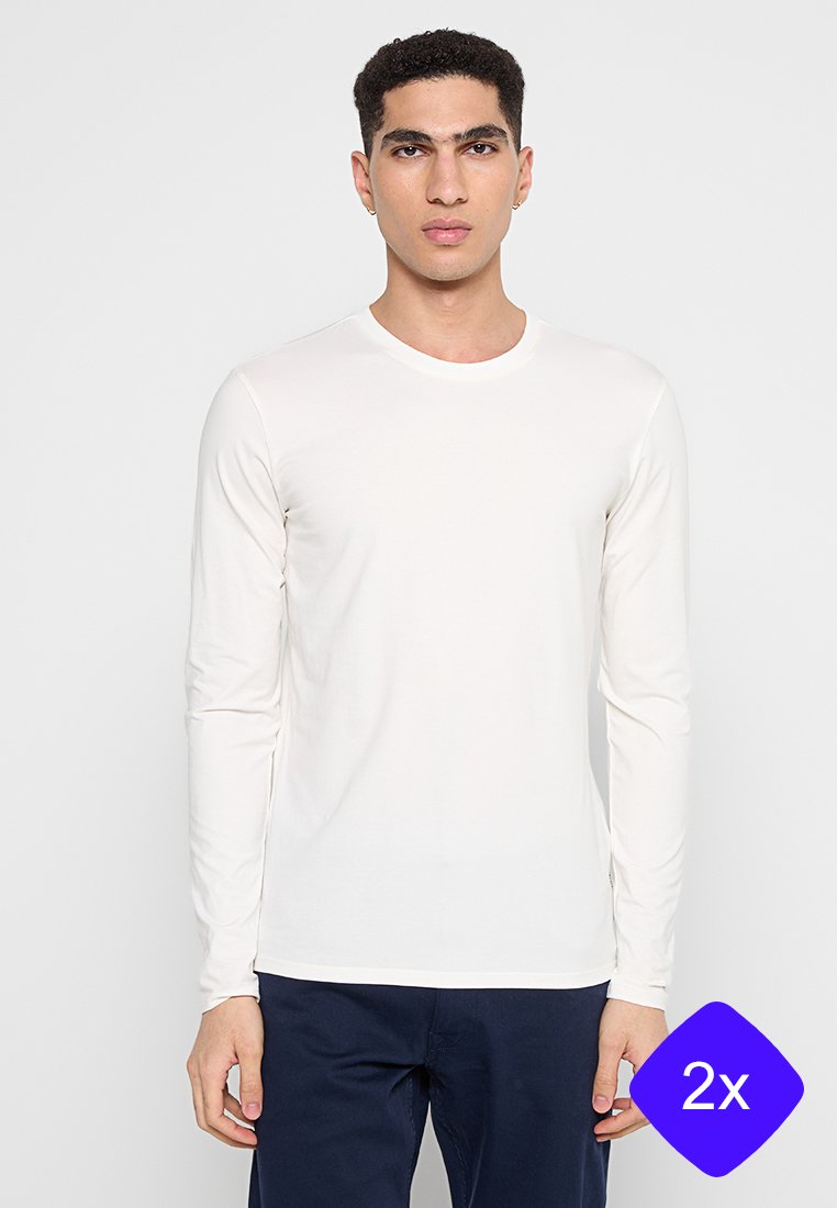 Biała koszula z długim rękawem, uszyta z miękkiego materiału, z okrągłym dekoltem, noszona z ciemnoniebieskimi spodniami. Charakteryzuje się czystym, prostym wzorem.