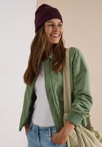 Femme portant un bonnet bordeaux, une veste verte, une chemise blanche et un jean bleu, souriante et portant un sac fourre-tout beige sur l'épaule.