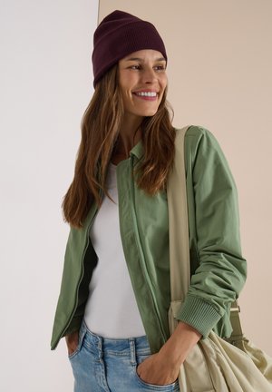 Frau mit dunkelroter Mütze, grüner Jacke, weißem Hemd und blauen Jeans, lächelnd und eine beige Umhängetasche über der Schulter tragend.