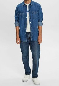 Camisa de denim en azul oscuro con botones en la parte delantera, dos bolsillos en el pecho y mangas arremangadas, combinada con jeans de azul claro y zapatillas blancas.