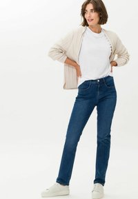 Blaue Jeans mit schmaler Passform, hoher Taille und fünf Taschen, kombiniert mit einem weißen T-Shirt und einem cremefarbenen, gerippten Cardigan. Weiße Sneakers runden den Look ab.