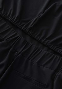 Zwarte stoffen jumpsuit met een elastische tailleband, subtiele textuur en een enkele achterzak. Glad, lichtgewicht materiaal.