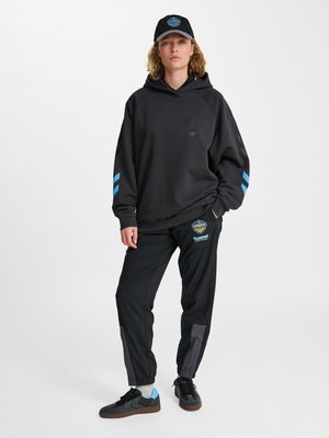 Młoda osoba nosi czarną bluzę z kapturem z niebieskimi paskami, czarne spodnie dresowe z logo, czarną czapkę i czarne sneakersy na białym tle.