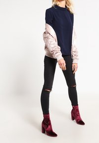 Felpa navy con collo alto, giacca bomber imbottita rosa chiaro, jeans skinny neri strappati e stivaletti in velluto bordeaux con tacco a blocco.