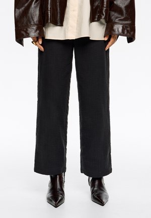 Pantalones negros texturizados con corte recto, combinados con botas de cuero negras de punta y una chaqueta marrón brillante de gran tamaño.