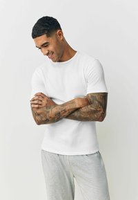 White ribbed short-sleeve t-shirt with a round neckline, paired with light grey sweatpants, on a person with tattooed arms.