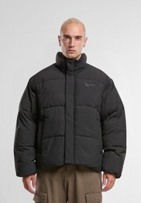 Homme avec des cheveux blonds courts portant une veste matelassée noire et un pantalon cargo marron sur un fond clair uni.