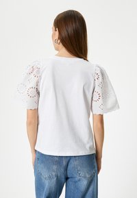 Hvid bomulds T-shirt med broderede, bølgede blonder puffærmer. Bæres med blå højtaljede jeans og et enkelt design.
