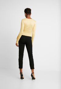 Enganliegendes gelbes Langarmoberteil kombiniert mit schwarzen maßgeschneiderten Hosen. Das Oberteil hat einen runden Halsausschnitt und die Hosen haben ein verkürztes Design.