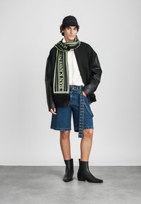 Chaqueta bomber negra, camisa blanca, pantalones cortos de mezclilla con acentos metálicos, bufanda verde y negra con texto, botas negras hasta el tobillo, fondo gris.