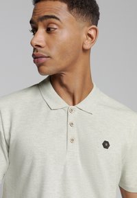 Lichtgroene polo met een kraag, drie knopen en een hexagonaal zwart embleem. De stof toont een geweven textuur voor extra detail.