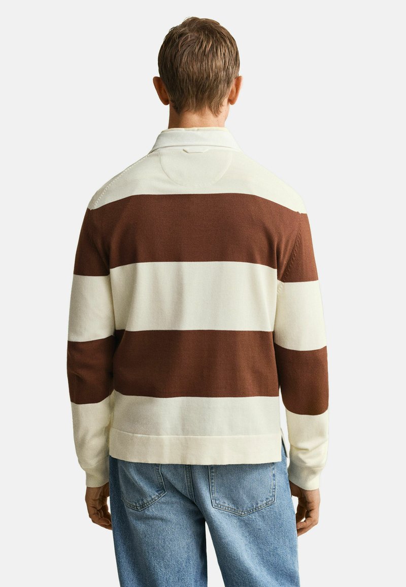 Mann trägt ein langärmeliges Polohemd mit breiten horizontalen braunen und cremefarbenen Streifen, kombiniert mit hellblauen Jeans, Ansicht von hinten.