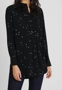 Chemise noire boutonnée ornée d'une broderie étoilée argentée, manches longues, encolure sans col et coupe décontractée.