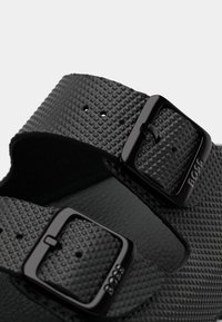 Gros plan sur des straps de sandales noires texturées avec deux boucles carrées noires estampillées du logo "BOSS".